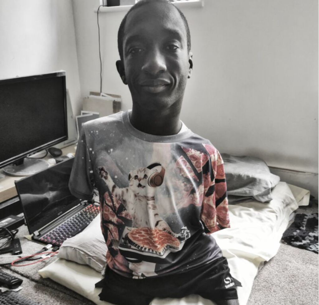 Photo of Isaac Harvey, a smiling young Black man. His tshirt has an image of a kitten wearing headphones and DJing with a pepperoni pizza. 
