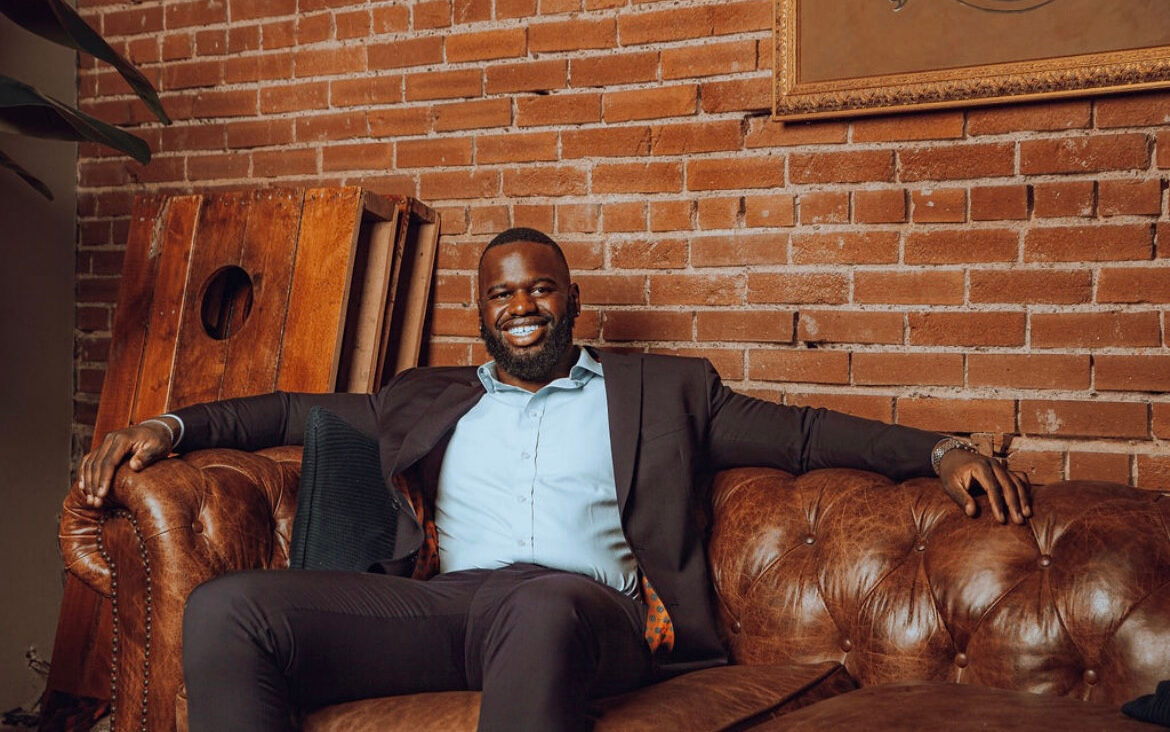 Solo Ceesay, a Black man with short hair, wearing a dark suit, sitting on a brown leather sofa smiling.