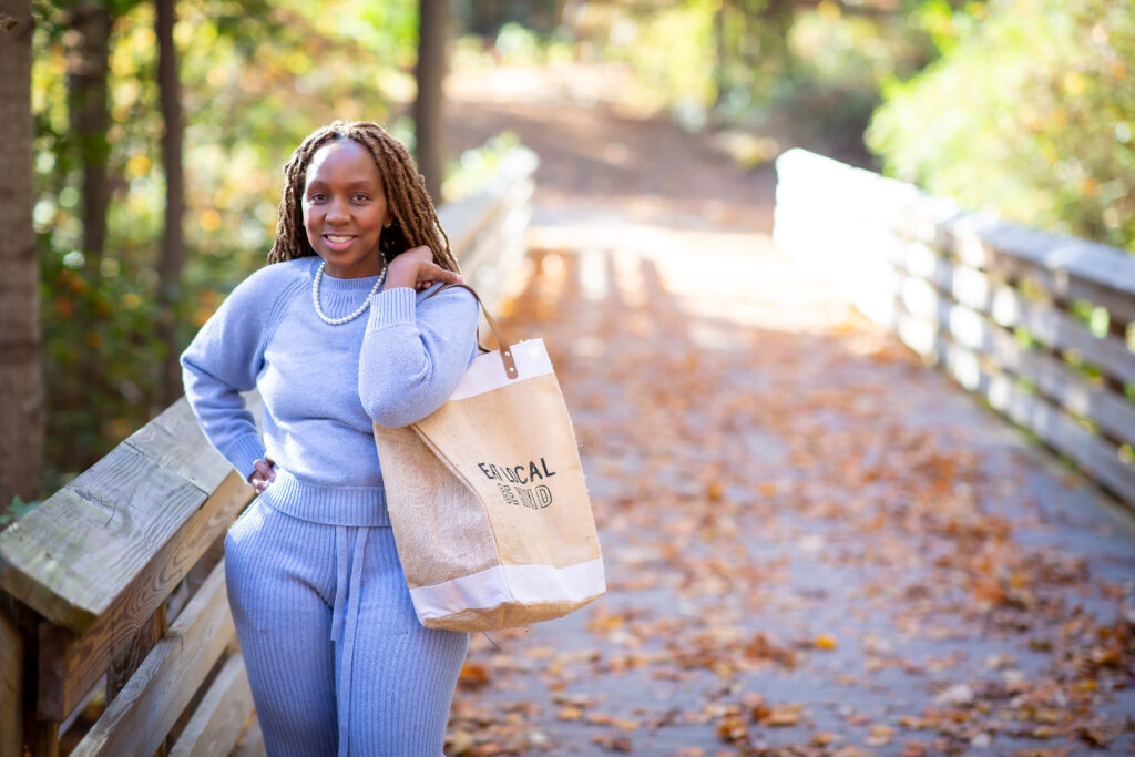 Photo of Eraina, a Black woman with long brown locs, standing on a woodland bridge smiling. She is wearing a purple top and trousers and carrying a canvas bag that reads 'Eat Local, Be Kind'