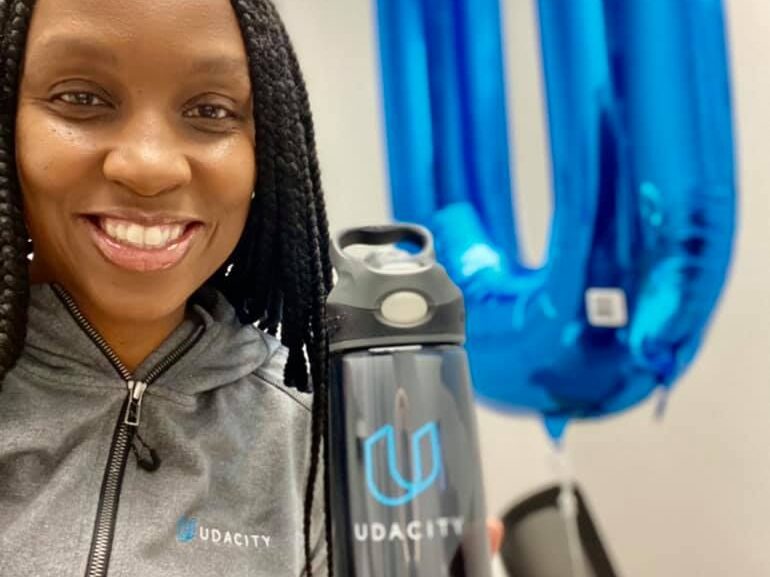 Photo of Eraina, a Black woman with long black braids, smiling at the camera holding a water bottle with the Udacity logo. She is wearing a grey hoodie with the Udacity logo