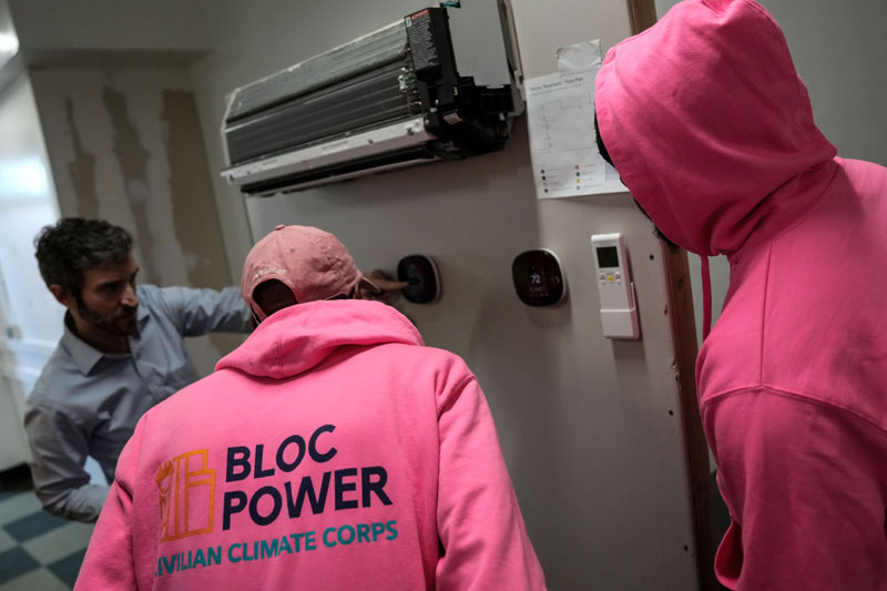 3 people looking at a device on the wall. Two are wearing pink sweatshirts with text that reads 'Bloc Power Civilian Climate Corps'