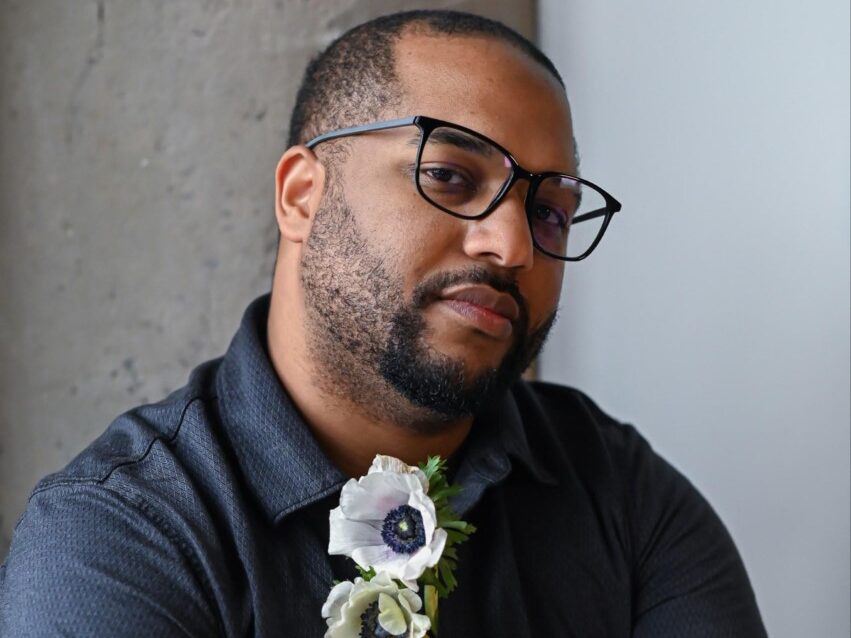 Black man with a beard wearing glasses and flowers in his shirt collar